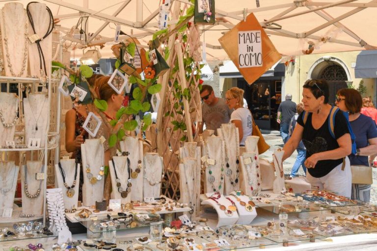 Como, Lake Como, Italy - June 2019: People looking at goods on a jewellery stall in the outdoor market in Como on Lake Como. A "Made in Como" sign is hanging from the stall